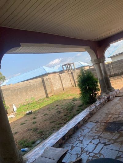 This media file depicts an outdoor scene in Jauro Yinu, Nigeria. The image is taken from a low-angle perspective, looking upwards and outwards from a covered area. The foreground features a section of a building's exterior wall with horizontal wooden paneling and a dark red trim.
Beyond this, an arched opening reveals the main scene. Two substantial concrete pillars, decorated with mosaic tiles near their tops, support the structure. Between the pillars, a lush green bush or small tree is visible, adding a touch of nature to the built environment.
In the mid-ground and background, a building under construction is evident. It is made of concrete blocks and has a partially completed structure covered with a light blue tarp or sheeting. The building appears to be multi-story with visible scaffolding or framework. The ground around the construction is dirt with patches of grass.
The sky above is blue with scattered white clouds, suggesting daytime with pleasant weather. The lighting indicates it is likely late morning or early afternoon. There are no people visible in the frame. A blue plastic bottle is placed on the ground near the tiled wall on the right. The overall impression is one of ongoing development in a semi-urban or rural setting in Nigeria.