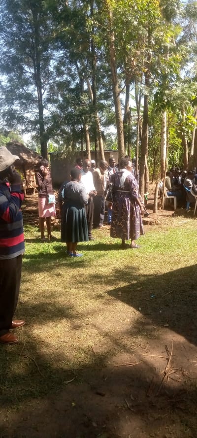The image shows a group of people standing in a grassy area. There are several trees in the background, some with large trunks.  The ground is covered in grass and scattered leaves. In the foreground, there are 11 people standing on the ground.  One of the people is wearing a red and white striped shirt and brown pants, and is standing towards the left of the image.  Another person is wearing a pink dress and is standing towards the left of the image. A third person is wearing a purple and black dress and is standing towards the center of the image. Several others are wearing casual clothing and are standing in a group near the center of the image. The scene is located in the town of Malava, Kenya. The coordinates are latitude: 0.508, longitude: 34.791.