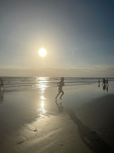 In Kuta, Indonesia, a child is silhouetted against the setting sun as they run along the wet sand of the beach. The sun, a bright orb, is low in the sky, casting a golden glow across the scene and reflecting off the water. In the background, other beachgoers are visible, some wading in the shallow water, while others are further out in the ocean. The waves are gentle, lapping at the shore, and the sand is marked with the footprints of those who have walked there. The overall atmosphere is one of peaceful enjoyment of a beautiful day at the beach.
