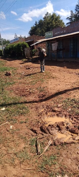 The image shows a dirt road in Lwandeti, Kenya. The road is muddy and there are puddles of water on it. There are buildings on either side of the road, including a small shop. A man in a blue shirt and jeans is walking on the road. The man has a backpack on his back. The scene is lit by the sun. The sky is blue with a few white clouds. There are power lines running overhead.
