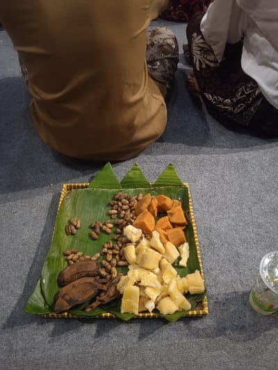 The image shows a plate of food sitting on a gray tiled floor. The plate contains a variety of food items, including sweet potatoes, bananas, cassava, and peanuts. Two people are sitting on the floor, but only their backs are visible. The scene is located in Surakarta, Indonesia, at coordinates -7.559 latitude and 110.783 longitude. The food is likely being prepared for a religious ceremony or a special occasion.