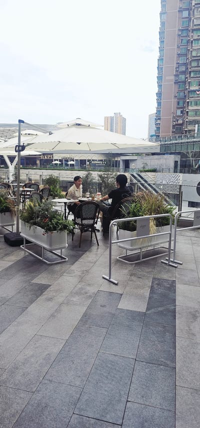 This image captures an outdoor cafe or dining area situated within a modern urban environment, likely a pedestrian plaza in Jiulongpo District, China. In the central foreground, two individuals are seated at a round table, occupying black and white patterned chairs. One person, dressed in a black t-shirt, is looking downwards, while the other, wearing a light-colored shirt, faces them with a slight smile, suggesting a pleasant conversation or shared moment.

The cafe area is equipped with several additional empty tables and chairs, all shaded by large, pristine white outdoor umbrellas. Planters filled with lush greenery, including ferns and other plants, are placed around the seating, creating natural dividers and adding to the ambiance. The ground beneath is paved with large, rectangular grey tiles of varying shades, arranged in a distinctive pattern.

In the mid-ground, behind the cafe, stands a modern building with a facade incorporating large glass panels and a visible escalator system. Further back and to the left, a tall residential building with numerous windows and air conditioning units is prominent. The background also reveals other distant high-rise structures under a bright, hazy sky.

Visible text includes "Sonny" and accompanying Chinese characters on the facade of a building, likely indicating a business or establishment. Partially visible text, possibly "chase" or a similar word, can be seen on the white metal barriers bordering the cafe area. The overall scene conveys a calm, daytime atmosphere, possibly late morning or early afternoon, characterized by soft, diffused lighting.