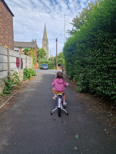 A young child wearing a pink hooded sweatshirt and yellow shorts rides a white bicycle with training wheels down a paved alleyway. The child's back is to the camera. To the left of the alley is a concrete wall with graffiti, followed by a brick building. To the right of the alley is a dense green hedge. In the background, a grey car is parked on a road, and a tall church steeple is visible above the trees. The sky is partly cloudy. The scene appears to be in a residential area of Salford, United Kingdom.