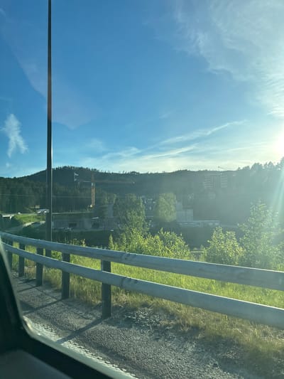 The image shows a view from a vehicle, looking out towards a hillside with buildings and construction. A tall, dark pole dominates the left side of the frame, obscuring part of the sky. The sky is a bright blue with wispy white clouds and a strong sun flare on the right. In the middle ground, there is a body of water or a lower elevation area with some structures, including what appears to be a tunnel entrance. A large orange construction crane is visible on the hillside. Further up the hill, there are several multi-story buildings nestled amongst trees. In the foreground, there is a metal guardrail and a roadside with gravel and grass. The overall impression is a semi-urban or industrial area on the outskirts of a town, likely Kongsberg, Norway, given the context. The scene appears to be captured during daylight.