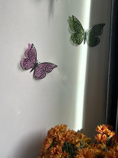 The image shows two decorative butterflies on a white wall. One butterfly is pink with intricate black filigree wings, and the other is green with similar black filigree. Below the butterflies and in the foreground, a bouquet of orange chrysanthemums is visible. A dark curtain is present on the right side of the image, and a vertical shaft of light illuminates the wall between the butterflies and the curtain. The scene appears to be indoors, likely in a residential setting, with no indication of Kathu, Thailand. There are no people or any actions depicted.