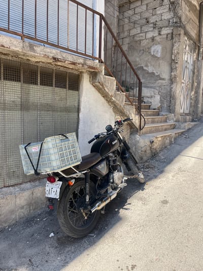 A black motorcycle with a rear delivery basket is parked on a street in what appears to be the As-Salihiya Municipality, Syria. The motorcycle is chained. The surroundings consist of concrete stairs, aged buildings, and a corrugated metal covering. The scene appears to be in an urban area. No people are visible.
