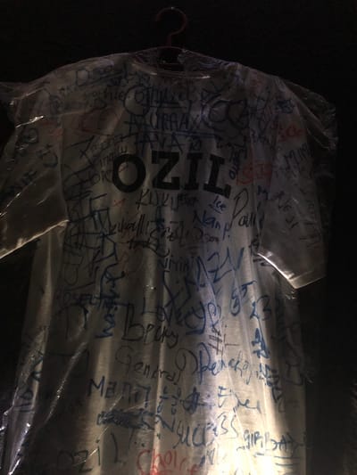 This media file shows a white t-shirt wrapped in plastic, covered in numerous handwritten signatures and messages in blue and red ink. The t-shirt is hanging from a hanger. The writing is dense and covers most of the visible surface of the shirt. Some of the visible text includes "OZIO", "MEHT", "Paul", "ice", "General", and various other names and scribbles.
The image appears to have been taken indoors or in a dimly lit area, possibly at night, given the dark background and the way light is reflecting off the plastic. There are no clear indications of weather or specific location cues beyond the provided context of "New Stadium, Nigeria." The overall impression is that this is a commemorative item, likely a t-shirt signed by attendees or participants at an event at the stadium. The focus is on the object and the inscriptions, with no people or specific activities clearly depicted.