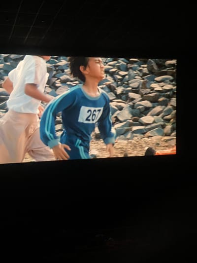 The image shows two individuals in athletic attire running outdoors, with a rocky terrain in the background. The person in the foreground, wearing a blue tracksuit with white and blue stripes, has the number "267" on their chest. The person behind them is wearing a white t-shirt and light-colored pants. The setting appears to be a daytime outdoor scene, possibly a race or athletic training event. There are no discernible elements within the image that would indicate the specific location in Banjarbaru, Indonesia.