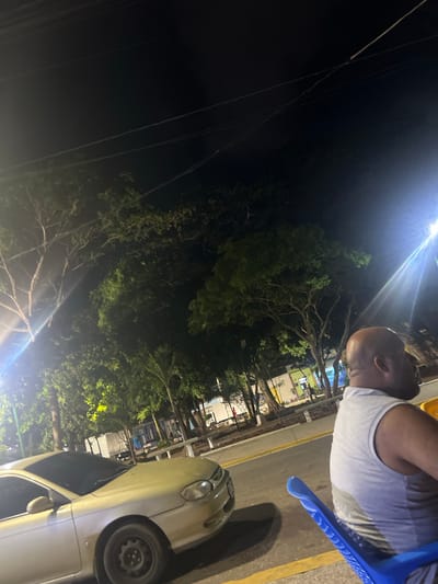 An outdoor night scene is depicted in El Valle del Espíritu Santo, Venezuela. In the foreground on the right, a bald man wearing a grey tank top is seated in a blue plastic chair. His body is angled away from the camera, looking towards the left. To the left of the man, a light-colored sedan is parked. The background features trees, streetlights casting bright beams, and what appears to be a street or plaza with buildings visible in the distance. Overhead, power lines are stretched across the dark sky. The lighting suggests it is nighttime, possibly in a well-lit urban or semi-urban area.