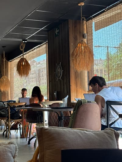 This image shows the interior of a cafe or restaurant, likely in València, Spain, with a focus on people seated at tables and the surrounding decor. The setting appears to be a space with a sloped ceiling covered in bamboo blinds, allowing natural light to filter through. Wooden panels line the walls and ceiling, creating a warm and rustic ambiance.
In the foreground, a person is seen from the back, seated at a table with a laptop. Further into the scene, another person, a young man, is bent over a laptop, wearing earbuds. He is wearing a white t-shirt and appears focused on his work. To his right, a person with long, dark hair, possibly a woman, is also seated, looking towards a screen. Another man with a beard is visible in the background, also looking down.
The cafe has unique lighting fixtures made of straw or similar material, resembling brooms, suspended from ropes. The furniture includes various chairs, some with brown padded seats and backs, and others made of lighter wood. The overall atmosphere suggests a relaxed daytime setting, possibly late morning or early afternoon, given the bright natural light. The presence of people working on laptops and enjoying the space indicates it's a functional cafe environment.