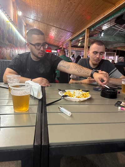 This image captures two adult males seated at an outdoor or semi-enclosed dining area in the city of Lom, Bulgaria, during evening or nighttime hours, indicated by artificial lighting. The ceiling structure above is constructed from wooden planks supported by beams.
In the foreground, two males are positioned at a beige, slatted table bisected by a dark central line. The male on the left has short dark hair, a beard, and wears black-rimmed glasses and a black patterned short-sleeved shirt. He is looking downwards and to his right. Two clear plastic cups containing a light amber liquid, consistent with beer, are placed directly in front of him. To his right, a white stack of paper napkins is held within a metallic dispenser. His left arm rests on the table, while his right arm is not fully visible.
The male on the right has short dark hair, a goatee, and wears a grey and black short-sleeved shirt. His left arm is extensively tattooed with intricate script and designs, and he wears a black smartwatch on his left wrist. He is actively engaged in placing a lit cigarette into a black ashtray with his right hand. His left hand holds a small object near his mouth, suggesting he is eating. A white plastic plate with french fries topped with crumbled white cheese and a black plastic fork rests on the table in front of him. Next to the ashtray, there is a blue pack of cigarettes (partially visible text includes Cyrillic characters), a white electronic cigarette or vape pen, a black lighter, and a red and white pack of "Omé" brand cigarettes.
The background to the left features a decorative wall with colorful murals and a wall-mounted lantern emitting a warm light. The background to the right shows blurred figures of other patrons and several overhead banners or awnings with white, inverted Cyrillic text, including what appears to be part of "ОБСЛУЖВАНЕ НА САМООБСЛУЖВАНЕ" (Self-Service), suggesting a casual dining establishment. A faint red glow from a distant sign is visible further back.