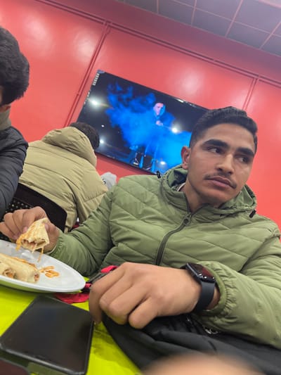 The scene depicts the interior of a casual eatery in Rukn ad-Din Municipality, Syria. A young adult male, with short dark hair and a light mustache, is positioned in the foreground, seated at a lime-green tabletop. He wears an olive-green quilted puffer jacket and a dark smartwatch on his left wrist. His gaze is directed towards the right side of the frame, away from the camera. His right hand holds a partially consumed wrap sandwich, revealing a light-colored filling. On a white plate to his left, additional wrap pieces and scattered orange-reddish crispy fragments are visible. A black smartphone lies face down on the table near his left hand, which rests on a dark bag or fabric item.
To the left of the primary subject, the shoulder and partial head of another individual in a dark jacket are visible. Further in the background, a third person is seated with their back to the camera, wearing a light tan or beige quilted puffer jacket. This individual is seated in a chair with a dark, perforated backrest. The background wall is painted a vibrant red, featuring horizontal and diagonal light-colored piping. A large flat-screen television is mounted on this wall, displaying an image with intense blue illumination surrounding a dark, humanoid figure. The overall lighting is artificial.