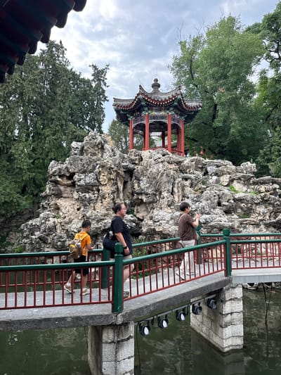 In Xicheng District, China, a bridge crosses over tranquil water, leading towards a rocky incline adorned with a traditional Chinese pavilion. Three individuals are present: a young boy in an orange shirt with a backpack, a woman in a black t-shirt with her hair in a bun, and another woman in a patterned shirt. The boy walks ahead, looking at his phone, while the woman in black walks alongside him. The woman in the patterned shirt is further ahead, holding a phone to her face as if taking a picture. The environment is lush with trees, and the sky is overcast, suggesting a cloudy day. The bridge features a red railing with green accents.