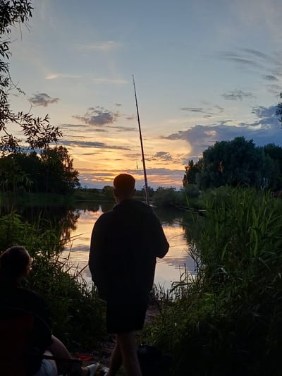 The image depicts a serene sunset scene by a body of water, likely a river or lake, with a person fishing. The sky is painted with warm hues of orange, yellow, and pink, indicative of a sunset. Clouds are scattered across the sky, reflecting the vibrant colors of the setting sun.

A person, silhouetted against the colorful sky, is positioned with their back to the camera, holding a fishing rod. The rod extends upwards and to the left, its tip subtly visible against the sky. The person is wearing dark clothing.

The immediate foreground is occupied by lush green vegetation, tall grasses and some trees, framing the water's edge. The water itself mirrors the sky, creating a beautiful reflection of the sunset and the surrounding greenery. Another person is partially visible in the lower right corner of the image, also facing away from the camera. The overall mood is peaceful and tranquil, suggesting a relaxing evening spent fishing.

The setting appears to be natural, possibly a rural or semi-rural area, given the dense vegetation and the calm water. The time of day is clearly sunset. There is no visible text in the image. The context provided indicates the location is Riga, Latvia.