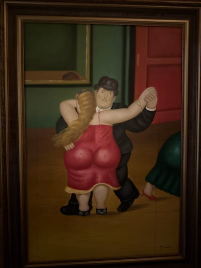 The image is a painting in a dark, ornate frame. The painting depicts two figures dancing. The figures are stylized with exaggerated roundness. A man in a black suit and hat is dancing with a woman in a red dress. The woman has long, blonde hair and her dress is adorned with small red polka dots and two larger red hearts on the bodice. Her arms are around the man's neck, and his arms are around her waist. Their hands are clasped above their heads. In the background, there is a green wall on the left and a red doorway on the right. There is also a glimpse of another figure in a green dress on the far right of the painting. The floor is a light brown wood. The painting is signed in the lower right corner with a cursive signature. The overall style of the painting is reminiscent of Fernando Botero's work. The image is a photograph of the painting, taken indoors.
This painting is located in the city of Heiligenrode, Germany.