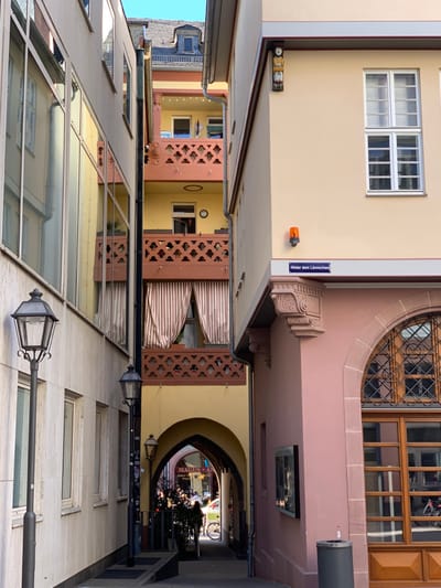 The image shows a narrow alleyway in Frankfurt am Main, Germany, located at 50.111 latitude, 8.683 longitude. The alleyway is bordered by two tall buildings. The building on the left is white with large windows and a street lamp. The building on the right is pale pink with a sign that reads "Hinter dem Lämmchen." A brown wooden door with a gridded window is visible on the right side of the building. A black trash can is placed next to the door. The alleyway is illuminated by sunlight, casting long shadows on the ground. There is a person walking in the distance towards an archway with a sign that reads "MAGUS - ART." There are a few other people visible in the distance, including a person riding a bicycle. An orange traffic light and a wooden figure are visible on the building to the right. The alleyway appears to be quiet and peaceful.