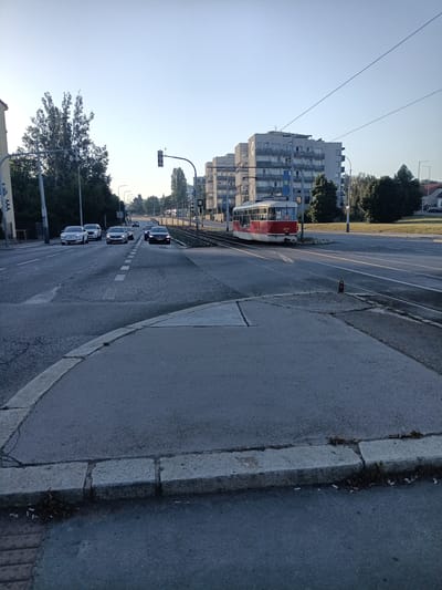 This media file depicts a street scene in Prague, Czechia, during what appears to be early morning or late afternoon due to the low angle of the sun casting long shadows. The sky is clear and blue.
The foreground shows a paved area with a concrete curb and scattered debris. A street intersection is visible, with a tram track running diagonally across the frame. In the middle ground, a red and white tram with the number "8777" displayed on its front is on the tracks, heading towards the right of the image. Several cars are driving on the road, including white, silver, and dark-colored vehicles, approaching from the distance. Traffic lights are visible at the intersection, indicating the flow of traffic.
The background features a row of multi-story apartment buildings with balconies, characteristic of urban residential architecture. Lush green trees line the street and surround the buildings. The overall setting is a typical urban environment with a mix of residential buildings, roads, and public transportation. The scene conveys a sense of normalcy and daily life in the city. There is no indication of any specific event taking place beyond typical urban traffic.