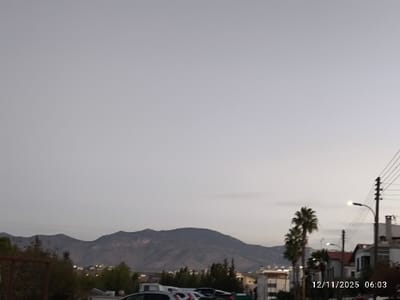A subdued cityscape unfolds beneath a vast, overcast sky. Rolling mountains loom in the background, their contours softly defined against the horizon. Palm trees and streetlights punctuate the urban landscape. Residences and parked vehicles are scattered throughout the scene. A timestamp marks the lower right corner. The overall atmosphere is muted, conveying a sense of tranquility.