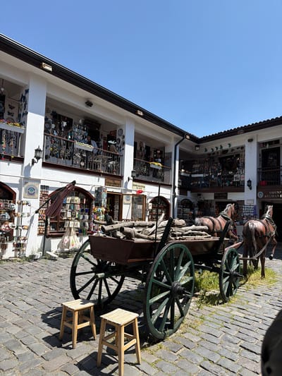 This media file depicts an outdoor scene in what appears to be a historic market or street, likely in Odunpazarı, Eskişehir, Turkey. The image is taken during the day under a clear, bright blue sky, indicating sunny weather.
The main subjects include a multi-story building with white walls and dark brown rooflines and balcony railings. This building houses several shops, with various crafts and goods displayed behind windows and on shelves, including ceramics, decorative plates, and other souvenirs. Some windows feature decorative iron grilles.
In the foreground, an antique-style horse-drawn cart is prominent. It is made of wood with large, dark green spoked wheels. Two lifelike, dark brown horse statues are attached to the cart, giving the impression they are pulling it. Beside the cart, two simple, light-colored wooden stools are placed on the cobblestone ground. The ground itself is paved with dark grey cobblestones, with sporadic patches of green grass growing between them.
There is no direct human activity visible, but the setting suggests a commercial or tourist area.
Visible text on shop signs includes:
- "ODUNP AZARI ATLIHAN E," which likely refers to a part of the Odunpazarı Atlıhan Handicrafts Bazaar.
- "YÜCEL LÜLETAŞI" (Yücel Meerschaum) with a phone number (234 59 48).
- "GİZEM SERAMİK" (Gizem Ceramics) with a phone number (0505- 232 29 69).
- A sign displaying "MAGNET."
- Partially visible text such as "NPAZARI" and "EDİYES."
The overall impression is one of a charming, traditional artisan quarter.