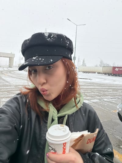 A female subject is photographed outdoors in a snowy environment, identified as the city of Argveta, Georgia. She is positioned in a medium-close-up, facing the camera with pursed lips. She has reddish-brown hair and is wearing a black newsboy-style cap, a light green hooded sweatshirt, and a black leather jacket. Snowflakes are visible on her cap and hair. In her right hand, she holds a white disposable coffee cup with a lid, branded "DUNKIN'", and a partially visible brown paper bag, also bearing "DUNKIN'" branding. Her visible fingernails are painted light pink with dark designs. The background features an overcast sky with active snowfall. The ground is paved and covered with a thin layer of snow, revealing vehicle tracks and yellow road markings. A tall street light pole is visible to the right. In the distance, a large concrete archway structure, multiple utility poles, and several semi-trailer trucks are present, one red trailer displaying "AS Logist".