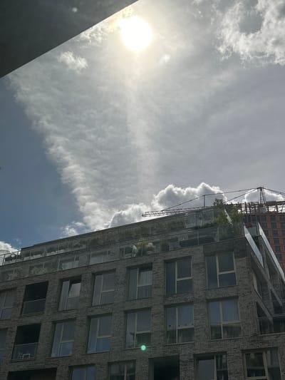 The image captures a sunny sky with scattered clouds above a modern multi-story building. The building features a gray brick facade with numerous windows and balconies. The rooftop appears to be a terrace with plants and glass railings. In the background, a construction crane is visible, suggesting ongoing development in the area. The scene is devoid of people. The lighting indicates it is daytime. The specific location in Amsterdam, Netherlands, is not identifiable from the visual information provided.