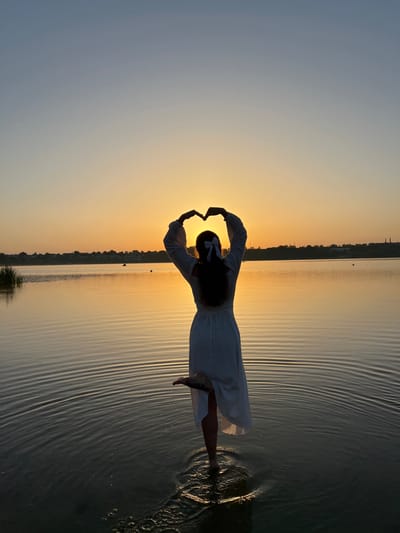 The image depicts a silhouette of a woman standing in shallow water at sunset. She is wearing a white dress and has her arms raised, forming a heart shape with her hands against the vibrant orange and yellow sky. The sun is partially visible, creating a warm glow that reflects on the calm water, which is rippled by gentle waves. A distant shoreline with vegetation and structures can be seen across the water. The overall scene is serene and picturesque, capturing a moment of peace and beauty at dusk. The location is Ternopil Lake in Ternopil, Ukraine, a popular recreational area.