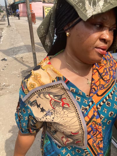 The scene depicts an adult female subject in a bright, outdoor urban environment identified as Umuode, Nigeria. The subject, positioned in the foreground and mid-shot, is a woman with dark braided hair partially covered by a dark green headscarf featuring a light-colored, possibly avian, design. She wears a multi-patterned garment, likely a blouse or dress, predominantly featuring teal, orange, beige, and black elements with equestrian motifs (stirrups, chains) and stylized facial patterns. She has a gold-colored stud earring in her visible right ear and a thin gold-colored chain necklace with a small cross pendant. Her head is turned slightly to the right, and her mouth is slightly ajar.
The background reveals a worn concrete sidewalk or pathway under strong daylight, casting shadows. A thin, dark pole stands behind the woman, and a reddish metal barrier is visible further down the path. In the distance, several buildings are present, including a light pink structure and another with a yellow sign displaying a partial "MTN" logo. A pedestrian wearing a blue backpack walks in the far distance on the left side of the path, with a faint glimpse of a small vehicle beyond. The overall environment suggests a bustling commercial or residential street.