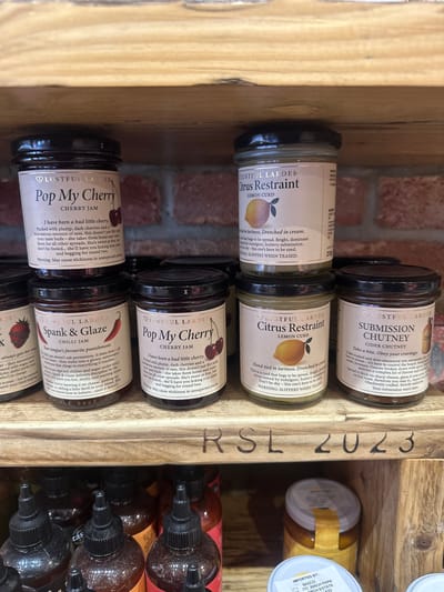 The image shows a wooden shelf in a shop, presumably in St Albans, United Kingdom, containing multiple jars of Lustful Larder artisanal condiments. There are visible jars of "Pop My Cherry" cherry jam, "Spank & Glaze" chilli jam, "Citrus Restraint" lemon curd, and "Submission Chutney" cider chutney. The jars are neatly arranged, showcasing the labels with descriptive text. A partially visible jar with a yellow label is also visible behind the Lusterful Larder products, and further back, a collection of bottles with black caps are also visible. The shelf itself is marked with "RSL 2023". A brick wall is visible in the background.