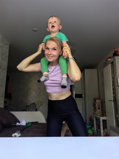 In the medium shot, a young woman with blonde hair is carrying a toddler on her shoulders indoors. The woman is smiling and looking at the camera. She is wearing a purple cropped t-shirt and black leggings. The toddler, wearing a green t-shirt and green leggings, is looking up with an open mouth, appearing excited. The child's feet are dangling down, one wearing a grey sock with an orange cartoon character. The background shows a living room with a couch, a wall with a patterned wallpaper, and a cabinet with shelves filled with items, including toys. The lighting suggests it is daytime, likely in the morning or afternoon. The scene conveys a sense of playfulness and joy between the mother and child.