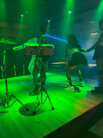 The image depicts a dimly lit nightclub or performance venue in Howayi, Nigeria, bathed in vibrant green stage lighting. In the foreground, a set of drum stands with cymbals and what appear to be bongo drums are visible. Three individuals are engaged in activity on the stage. To the left, a person is partially obscured, facing away from the camera and wearing white pants. In the center, a person with dark hair is dancing, wearing a white strapless top and a dark, short skirt, with white high-top sneakers. To the right, another person, also with dark hair, is visible from the back, wearing a dark skirt and dark shoes, with their hand extended. The stage floor is a glossy wooden surface reflecting the green lights. Several vertical, sword-like objects are placed along the back wall of the stage. The overall atmosphere suggests a live performance or entertainment event.