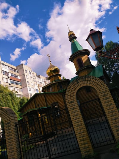 The image shows a wooden church with a golden dome and a green roof, situated in the city of Horishni Plavni, Ukraine. The church is framed by a brick archway and a black metal fence. In the background, there is a multi-story apartment building and lush green trees. The sky is blue with white clouds. There are no people visible in the image. The perspective is from a low angle, looking up towards the church and the sky. The lighting suggests it is daytime.
