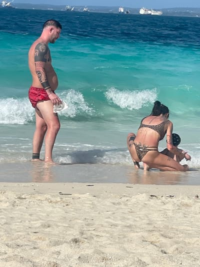 On a beach in Zanzibar, Tanzania, a male, a female, and a child are in shallow ocean water. The male, shirtless and with extensive black arm and torso tattoos, wears red patterned swim briefs. He stands barefoot in the ankle-deep turquoise water, holding a small white object in his right hand, looking downwards and slightly to his right. To his immediate right, a female with dark hair tied back, wearing a leopard print two-piece bikini and displaying multiple tattoos on her back and lower back, squats in the water. She faces away from the camera, attending to a small child, partially obscured by her, who wears a dark cap. Small waves break around the individuals. The immediate foreground shows light-colored sand. The ocean transitions from clear turquoise to a deeper blue further from the shore. Several large ships are visible on the distant horizon under a clear sky.
