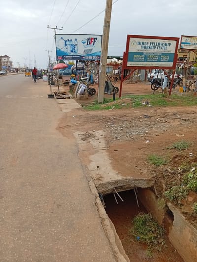 The image depicts a street scene in Sabon Tasha, Nigeria, near the Bible Fellowship Worship Centre at KM 10 Kachia Road. A damaged concrete drainage ditch is visible in the foreground, with exposed rebar and debris inside. A road runs alongside the ditch. Further down the street, a "POS" stand is set up near a bicycle repair area, and a man is walking. In the background, a large advertisement for DFF table water is visible.
