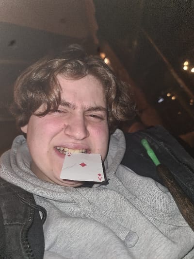 A person with curly brown hair and a fair complexion is shown with the Ace of Diamonds playing card stuck between their teeth. They are wearing a gray hoodie with a dark jacket partly visible in the frame. The lighting is dim, and the photo is slightly out of focus. The background is dark and blurry, with some faint light sources visible. The image appears to be taken from an unusual angle, possibly tilted.