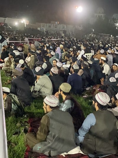 A large crowd of men sit outdoors at night in what appears to be Kandahar, Afghanistan. Many wear traditional pakol hats and some have beards. The scene is illuminated by bright lights, and a building can be seen in the background. The men are seated on grass and possibly rugs.