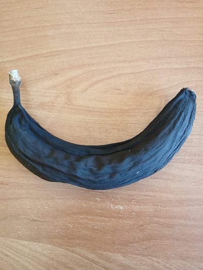 A very dark, dried banana lies on a light brown, wood-grained surface. The banana is curved, with its stem at the bottom left and the opposite end curving upwards and to the right. The peel is a deep black, with visible wrinkles and lines indicating it has dried out significantly. The stem end has a small, dry, fibrous tip. The wood surface has a fine grain and a few scattered light-colored specks, possibly dust. The lighting appears to be from above, casting a slight shadow beneath the banana, though the surface is generally evenly lit. There is no text visible in the image. The scene is simple and focused on the solitary, desiccated fruit against the textured background.