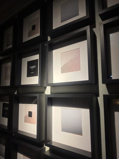 The image shows a wall with multiple framed artworks, likely a gallery or a cafe, in the city of Medan, Indonesia. The frames are dark, creating a contrast with the lighter matte borders and the artwork within. The artworks themselves are abstract and geometric, featuring patterns of dots, squares, and textured gradients. One frame contains text that reads ".TEMU café medan", indicating this is likely a cafe or restaurant named TEMU in Medan. The lighting in the scene is dramatic, with strong shadows cast across the artworks and the wall, suggesting directional spotlights or natural light entering from an angle. The overall impression is one of a modern and minimalist display of art.