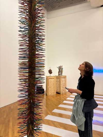 A woman stands in an art gallery, examining an art installation. The installation in the foreground consists of colored pencils protruding from a piece of wood. Behind this is another wooden installation with sticks and found objects. The woman is holding a smartphone, possibly taking a picture of the art. The gallery has a white wall and a striped floor. A neon sign is visible in the background. This art gallery is located in the city of São Paulo, Brazil.