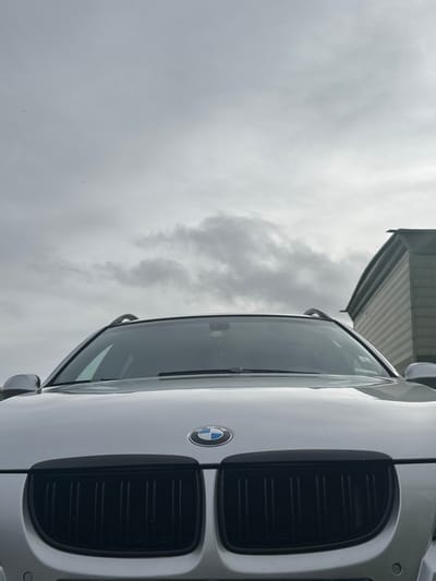 The image displays the front of a silver BMW vehicle, featuring black kidney grilles and a centered BMW emblem on the hood. Roof rails are visible on the vehicle's roof, above the windshield. The scene is outdoors in Muirhead, United Kingdom, under an overcast sky with gray clouds. To the right of the vehicle, a portion of a light-colored outbuilding with horizontal paneling and a sloped roof is present. No individuals or activities are depicted.