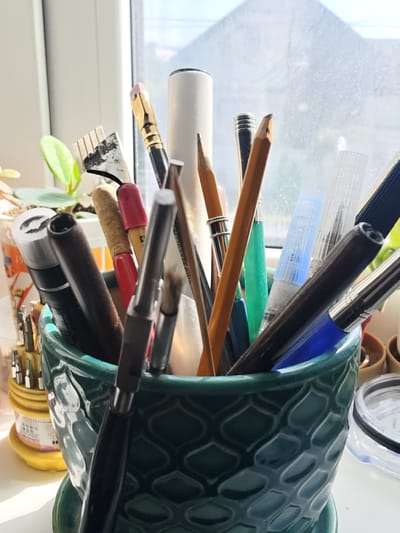 The image depicts an assortment of art supplies and tools arranged in a dark green, textured ceramic pot. The pot is filled with various items including pencils, brushes, pens, and what appears to be a small toolkit with multiple bits. Sunlight streams in from a window in the background, casting shadows and highlighting the textures of the objects. The window is partially obscured by the subjects, but a blurry view of what could be buildings or trees is visible outside. The items are densely packed, suggesting they are actively used. The setting appears to be a well-lit indoor space, likely a workspace or studio, with a plant visible to the left of the pot. There are no people present in the image. The scene is static and focuses on the organization of creative tools. The location is specified as Colonița, Moldova.