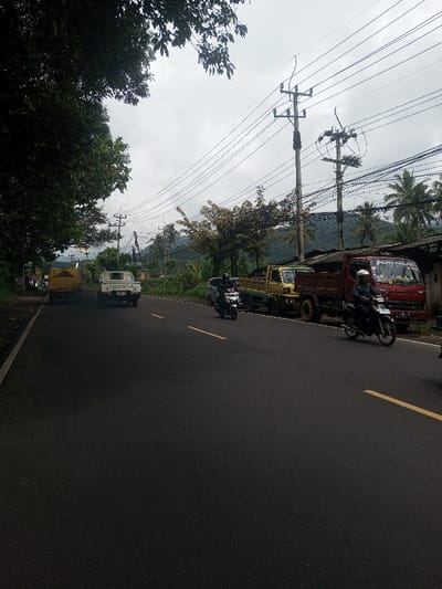 A rural road scene shows a yellow Toyota truck followed by a white Toyota pickup. A motorcycle rides in the same direction. Parked along the road are a yellow truck and a red dump truck. Another motorcycle is also on the road. Utility poles and wires cross overhead with trees and buildings along the roadside. Mountains are visible in the background under a cloudy sky.