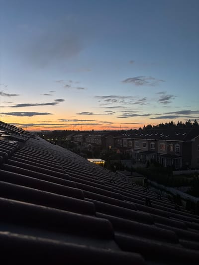 An elevated perspective captures a twilight scene over a residential area in Baranovo, Russia. The foreground is dominated by a dark, tiled roof, angled diagonally to the right, its texture and rows of tiles clearly defined in the dim light. Beyond the roof, a line of terraced houses stretches across the middle ground. These buildings are predominantly light-colored with dark roofs and evenly spaced windows, some of which are illuminated from within. A white picket fence runs along the front of the properties. Behind the houses, a dense line of dark green trees forms a backdrop, suggesting a wooded area. The sky above is a gradient of soft blues and grays, with streaks of orange and yellow near the horizon, indicating the setting sun. Scattered clouds, ranging from wispy to more substantial, drift across the sky. The overall impression is one of a quiet, suburban evening. There are no people or animals visible in the image.