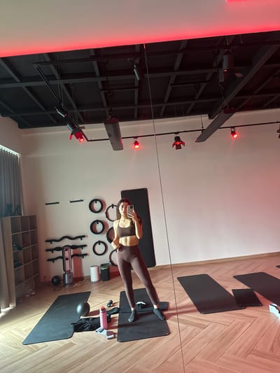 The media file shows a fitness studio with a woman in athletic wear taking a selfie. The studio features wooden flooring with a herringbone pattern, and several yoga mats are laid out. On the wall, there are various fitness accessories such as resistance rings, weights, and what appears to be a Pilates reformer. Red lights are strategically placed around the studio, casting a dramatic illumination. The woman is in a workout outfit, posing for the photo. The scene suggests an active or preparatory moment in a fitness session. The overall ambiance is modern and professional.