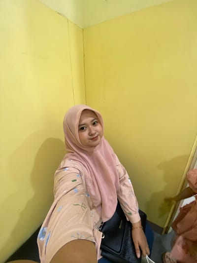 In Sidoarjo, Indonesia, a young woman wearing a pink hijab and a floral-patterned light pink blouse is captured in a selfie. She is seated, with her right arm extended, holding a white bag. Her left arm rests on a black handbag, which is placed on her lap. The background consists of a plain yellow wall. The lighting suggests an indoor setting, possibly a room. The image focuses on the woman and her immediate surroundings.