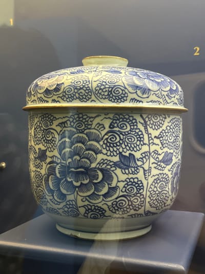 A blue and white ceramic jar, likely made of porcelain, is displayed in a museum or exhibition setting. The jar is horizontally oriented and features intricate underglaze cobalt blue floral decorations against a light, possibly white or cream-colored, background. The patterns include large, detailed peony-like blossoms, smaller swirling chrysanthemum-like motifs, and delicate foliate designs. A distinct, narrow band with a metallic or golden sheen runs horizontally around the middle of the jar, suggesting it might be a lidded vessel or constructed in sections. The object is seen against a muted blue-grey background, which appears to be a display case or wall. In the upper left background, a golden number "2" is visible, likely serving as an exhibition label. The lighting suggests an indoor environment. This artifact is located in Tepotzotlán, Mexico.