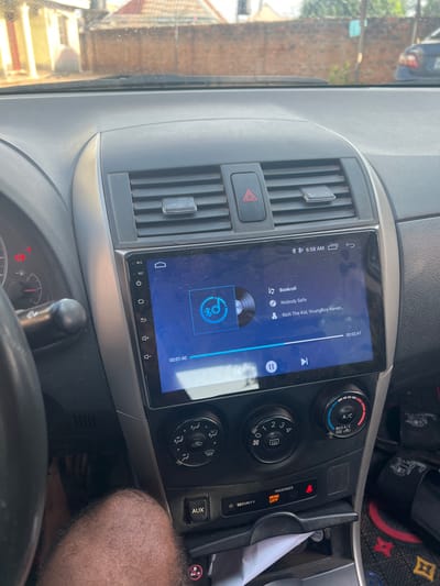 This image shows the dashboard of a car, specifically the center console area. The car is playing a song called "Nobody Safe" by Rich The Kid and YoungBoy Never Broke Again from the album "Bankroll" at 6:58 AM. The car's air conditioning and heating controls are visible, and the passenger airbag indicator is set to "off". There's a partial view of a person's hairy legs in the bottom right corner, along with a glimpse of the car's steering wheel. Based on the left side of the image, the car may be located in a city with greenery, possibly Markurdi, Nigeria.