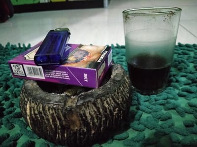 The image shows a glass of dark liquid, possibly coffee, on a green carpet, a blue lighter sitting on top of a pack of cigarettes, and a dark brown, circular ashtray. The pack of cigarettes is purple and white, and the front shows a barcode and the words "43 MG TAR, 2.2 MG NIKOTIN" in white font. The words "SKT" appear in white font on the bottom of the pack. The carpet has a soft, fuzzy texture. The blue lighter is in focus, and the glass of dark liquid and pack of cigarettes are slightly out of focus. The image was taken in a home or bedroom, most likely in the city of Lagadar, Indonesia, based on the provided coordinates. The scene suggests a casual setting where someone is enjoying a cigarette and a drink. The image does not show any people.