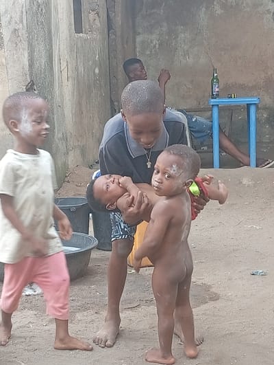 An outdoor scene in Owerri, Nigeria, depicts four children and one adult. In the foreground, from left to right: A young boy, approximately 4-6 years old, wearing a light t-shirt and pink cropped pants, walks right, looking towards the right and smiling. White powder is visible on his forehead and left cheek. His feet are bare. A slightly older child or teenager, approximately 10-14 years old, with short hair, wears a dark blue polo shirt with a light grey collar and patterned shorts. He has a metallic cross necklace. He holds an infant, approximately 6-12 months old, horizontally in his arms; the infant has white powder on its forehead and a red and green diaper on its right thigh, looking upwards. To the right, a toddler, approximately 1-2 years old, stands nude with white powder on its forehead and right cheek, looking directly at the viewer. A red cloth with a green band is held near its right leg. All children in the foreground are barefoot.

In the mid-ground, to the left, two dark-colored plastic basins are on the unpaved, dusty ground. In the background, to the right, an adult male is partially visible, seated on a low structure, wearing blue jeans and flip-flops, leaning against a textured, light brown concrete wall. A small, light blue plastic table stands next to him, holding a green glass bottle and a small yellow object. A rectangular opening is visible higher up in the wall. The ground consists of packed earth or sand.