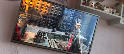 An indoor television screen displays a scene from a movie or show. The on-screen image depicts a woman in historical attire, including a hat and a long cloak over a pink dress with lace trim, standing on a metal grate floor. Behind her, a stone wall and a structure with many drawers are visible. A candle and a metal cage are present in the foreground of the on-screen scene. The television itself is mounted on a wall with textured pink wallpaper. Above the television, a shelf holds several decorative items, including ceramic pigs and a white bear figurine. The location is a room, likely a living room, in a residential setting. The city of Tikhvin, Russia, is not depicted in this image.