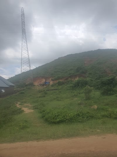The image depicts a rural scene on Rukuba Road, Nigeria, under a cloudy sky, suggesting a temperate or overcast day. The primary subjects are a large metal pylon and a blue truck on a dirt road that runs alongside a lush, green, and heavily vegetated hillside. The hillside itself appears to be a significant geological feature, possibly a mountain or large incline, with some areas showing exposed reddish-brown earth, indicating potential erosion or construction activity.
The pylon, a prominent man-made object, is situated in the foreground, extending towards the upper left of the frame. It is a lattice-type structure, indicative of power transmission lines. The truck, a utilitarian vehicle, is positioned on the dirt road higher up on the hillside, and there appears to be at least one person near it, possibly engaged in work related to the road or the power lines. The presence of both the pylon and the truck suggests human activity in this natural landscape, likely related to infrastructure development or maintenance.
The scene is characterized by the juxtaposition of industrial elements with the raw, natural environment. The greenery of the hillside is thick and appears to be largely untamed, while the dirt road and the pylon represent human intervention. The cloudy weather contributes to a subdued atmosphere, with soft, diffused light. There are no visible signs of text within the image. The overall impression is one of a functional landscape where human infrastructure is integrated into a rugged, natural setting.